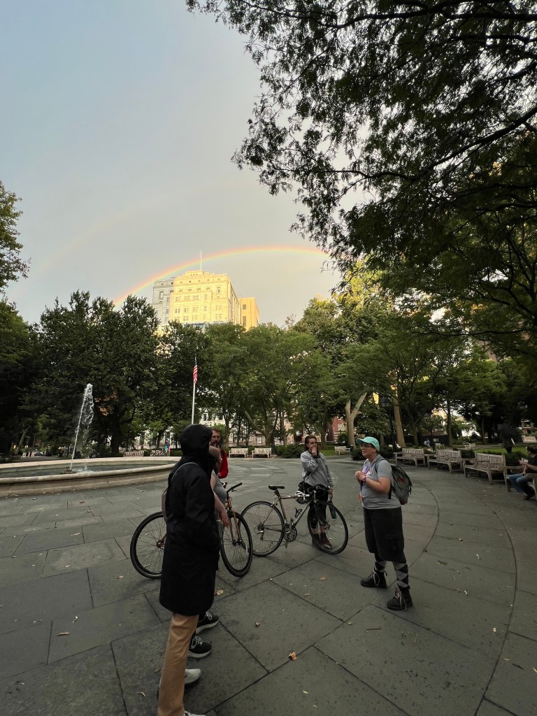 Tour guide speaks to a small group in Washington Square Park under a full rainbow